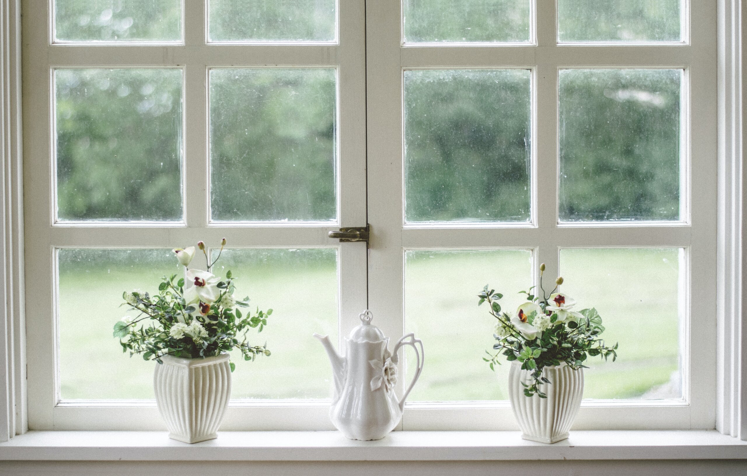 prozor s bijelom prozorskom klupčicom na kojoj su dvije bijele tegle s cvijećem i porculanski čajnik u sredini